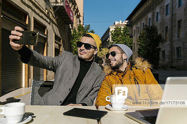 Junge schwule Männer mit Sonnenbrille  die ein Selfie mit ihrem Handy machen  während sie in einem Straßencafé sitzen