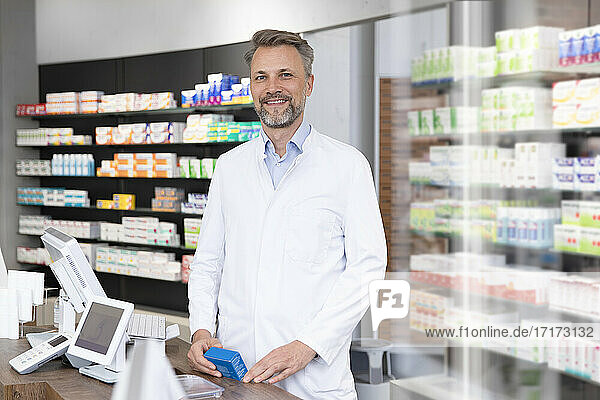 Smiling mature pharmacist with medicine box standing at pharmacy checkout