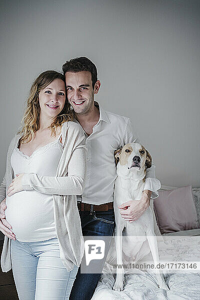 Lächelnder Mann mit schwangerer Frau und Hund im Schlafzimmer stehend