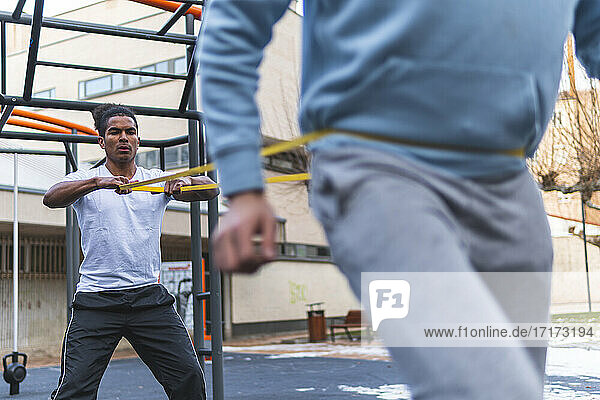 Young man holding stretching band while exercising with friend in city