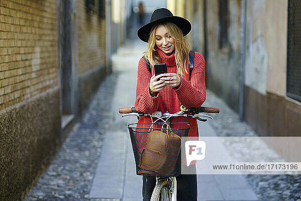 Blond woman using smart phone while standing with bicycle on footpath