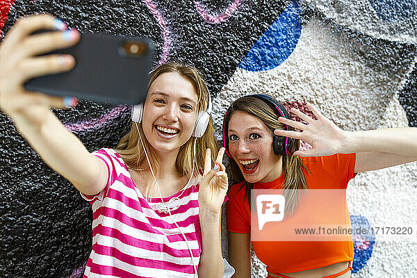 Happy female friends taking selfie through smart phone while gesturing against graffiti wall