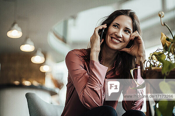 Lächelnde Geschäftsfrau  die wegschaut  während sie in einem Café mit einem Smartphone spricht