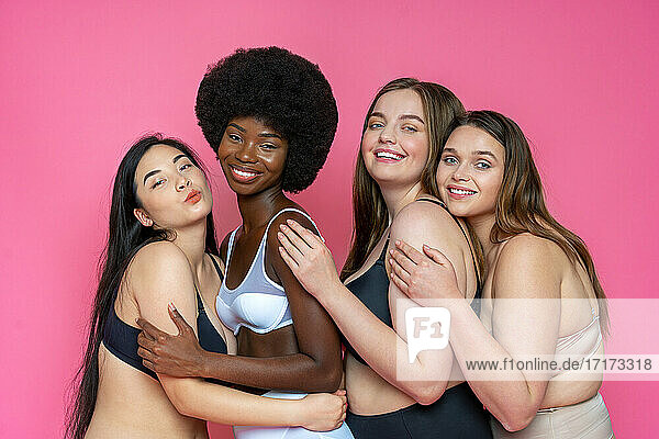 Smiling group of multi-ethnic female models in lingerie embracing each other against pink background