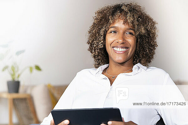 Lächelnde Frau mit digitalem Tablet  die wegschaut  während sie zu Hause sitzt