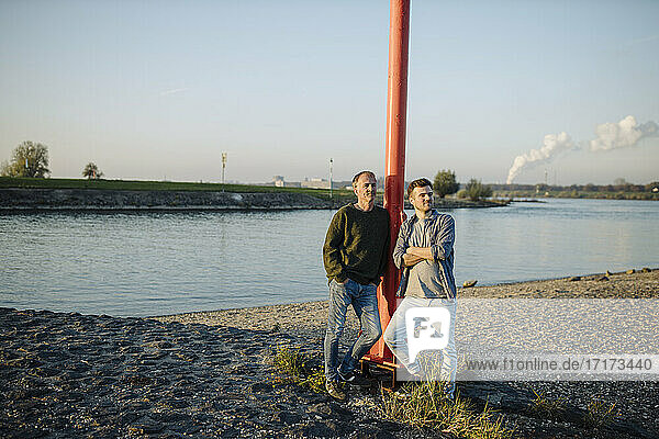 Thoughtful father and son leaning by pole against sky at riverbank