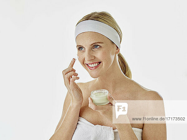 Young woman smiling while applying facial cream standing against white background
