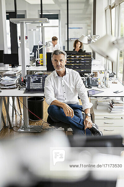 Mature businessman sitting on chair with colleague standing in background at open plan office
