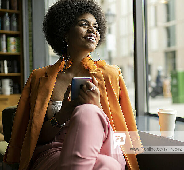 Smiling woman with smart phone looking out of window in cafe