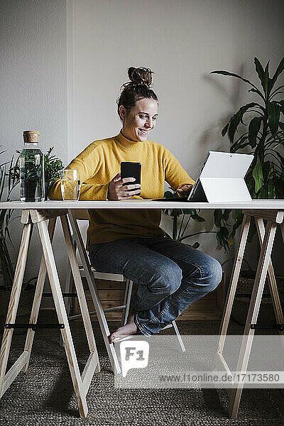 Smiling woman with mobile phone using digital tablet sitting at home