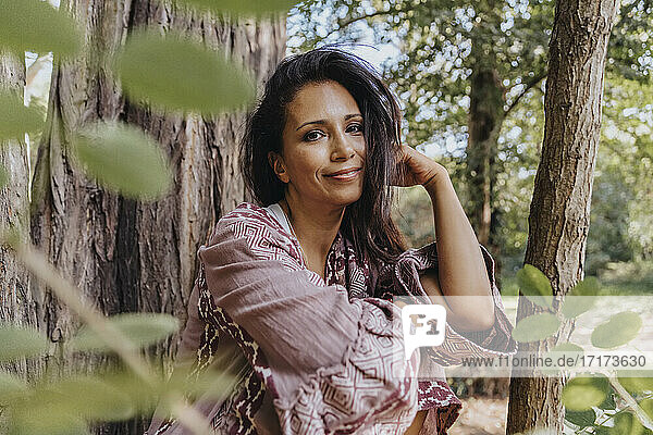 Mature woman with hand in hair smiling while sitting under tree