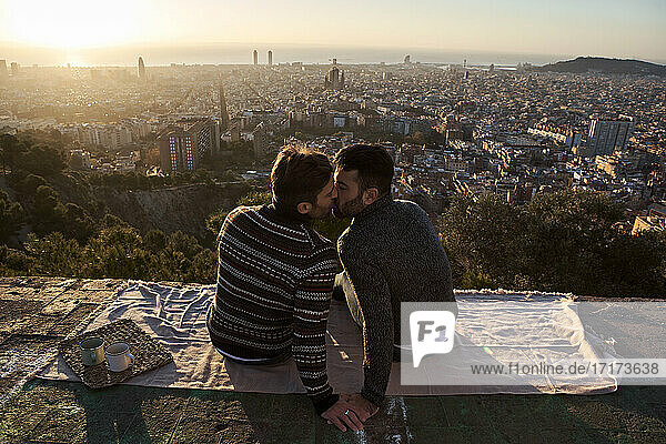 Schwule Männer  die sich küssen  während sie auf dem Aussichtspunkt vor der Stadtkulisse sitzen  Bunkers del Carmel  Barcelona  Spanien