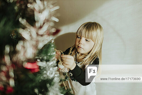 Blond girl decorating Christmas tree against wall at home