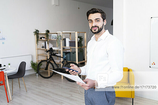 Lächelnde männliche Fachkraft mit Papierdokument  die ein Mobiltelefon bei der Arbeit am Arbeitsplatz hält