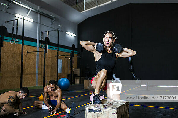 Female athlete lifting dumbbells while exercising on box and men resting in gym