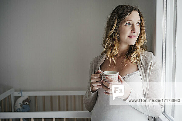 Thoughtful pregnant woman holding coffee mug while standing against crib at home