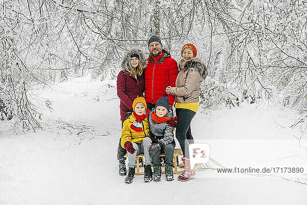 Smiling parents standing by children sitting sled in forest during winter