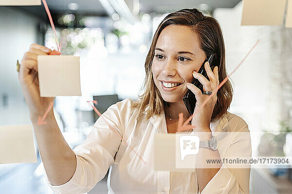Nahaufnahme einer lächelnden Geschäftsfrau  die über ein Mobiltelefon spricht  während sie auf einen auf Glas aufgeklebten Haftnotiz schreibt