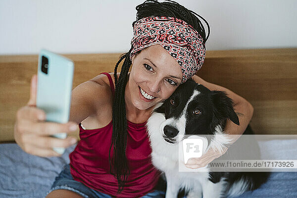 Smiling woman taking selfie with pet through mobile phone while sitting on bed at home