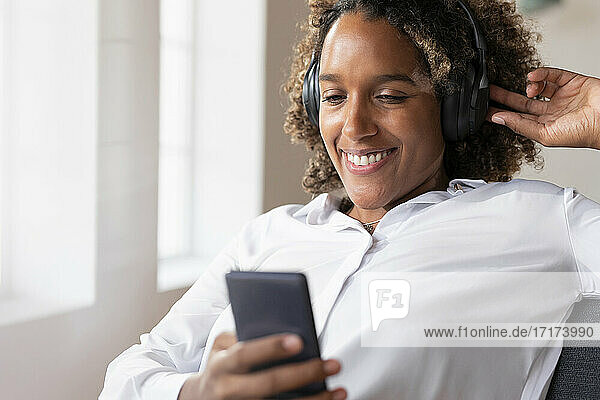 Lächelnde Frau  die Musik hört  während sie ein Mobiltelefon benutzt und zu Hause sitzt