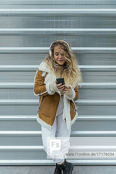 Young woman listening music through smart phone while standing against metal wall