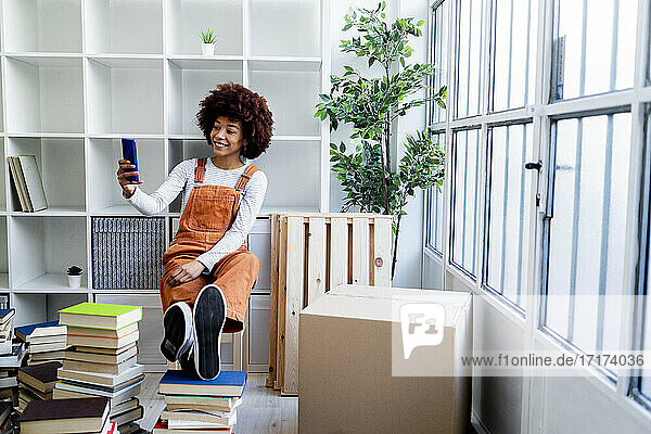 Lächelnde Afro-Frau  die ein Selfie vor einem Bücherregal macht  während sie in eine neue Loft-Wohnung zieht