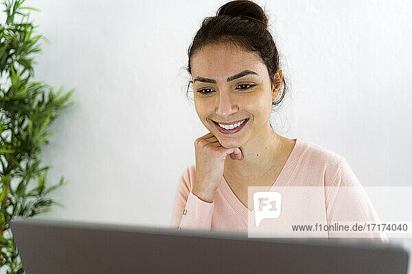 Smiling woman with hand on chin working on laptop while sitting at home