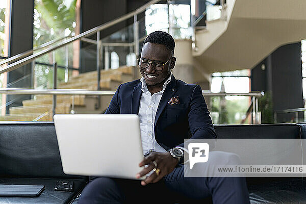 Lächelnder junger männlicher Unternehmer  der auf einem Sofa in einer Hotellobby sitzt und einen Laptop benutzt