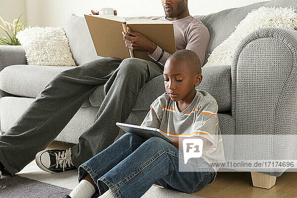 Junge sitzt auf dem Boden und benutzt ein digitales Tablet  Vater liest ein Buch