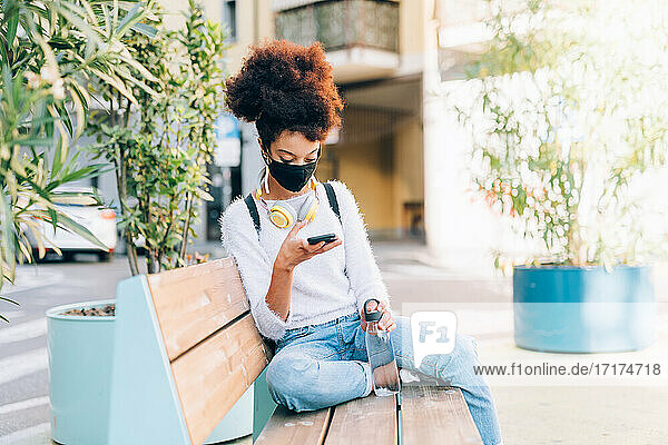 Junge Frau sitzt auf einer Bank  trägt eine Gesichtsmaske und schaut auf ein Telefon