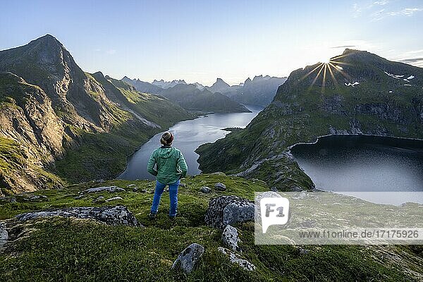 Junge Frau blickt über Fjordlandschaft  Sonne strahlt über Berglandschaft mit Fjord Forsfjorden und See Krokvatnet  Moskenesöy  Lofoten  Nordland  Norwegen  Europa