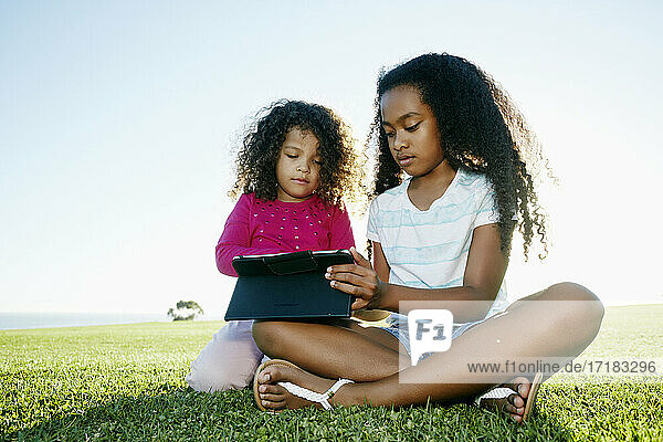 Junges gemischtrassiges Mädchen und ihre jüngere Schwester sitzen im Freien und teilen sich ein digitales Tablet
