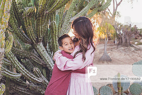 Mutter umarmt Sohn vor einem großen Kaktus mit Gegenlicht Sonne.