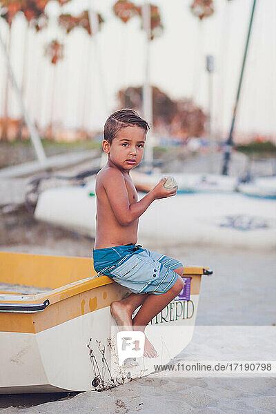 Young boy sitting on a docked boat at the beach and holding a ball.