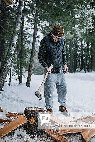 A man wearing beanie and flannel chops firewood in the snow