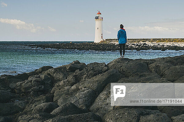 Junge Frau mit Blick auf einen Leuchtturm in der Ferne  der auf der felsigen Küste steht  Port Fairy  Australien.