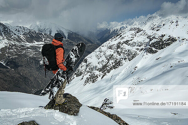 Mann mit Snowboard steht auf einem Felsen an einem schneebedeckten Berg gegen einen bewölkten Himmel