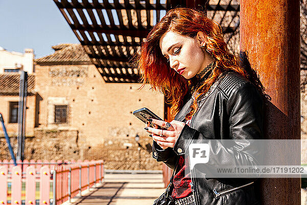 Alternatives Mädchen mit blauen Augen und orangefarbenen Haaren  das sich an eine Säule lehnt  während sie ihr Mobiltelefon benutzt.