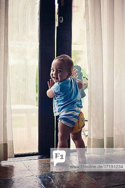Kleiner barfüßiger Junge hält sich an einer großen Glastür fest.