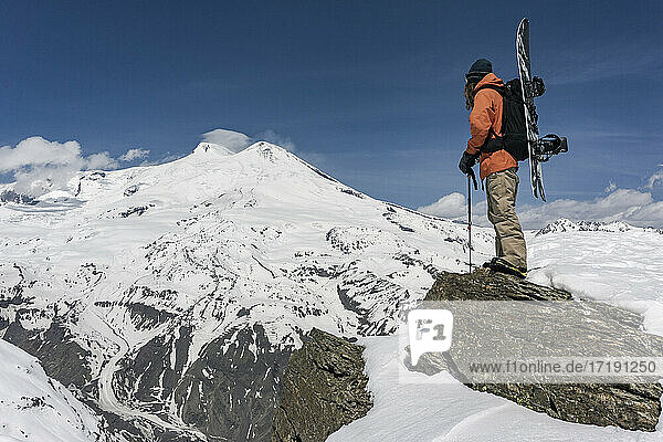 Mann mit Skistock und Snowboard steht auf einem Felsen vor einem schneebedeckten Berg gegen den Himmel