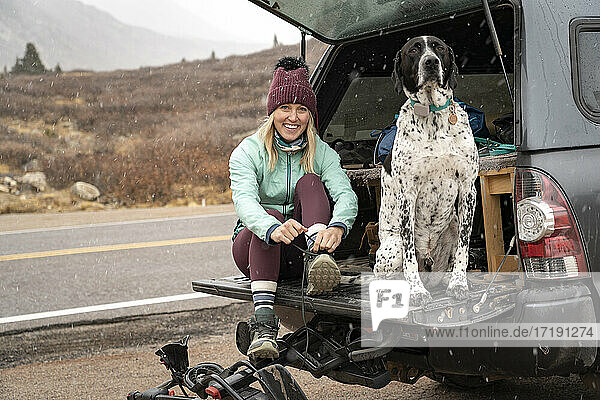 Porträt einer lächelnden jungen Frau  die mit ihrem Hund im Kofferraum eines Geländewagens sitzt und die Schnürsenkel bindet