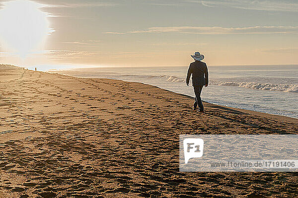 Mann  der mit einem Hut bei Sonnenuntergang am Strand spazieren geht