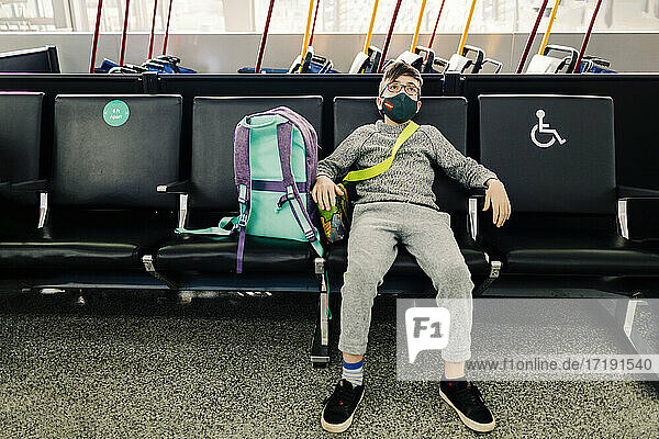 Ein Junge mit Brille und Maske sitzt im Flughafenterminal und wartet