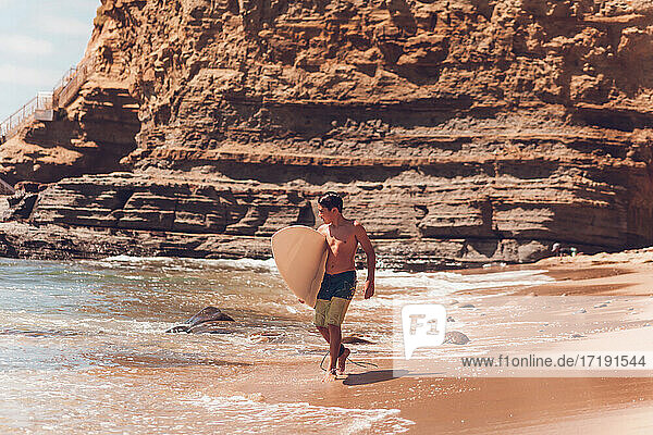 Junge  der am Strand spazieren geht und sein Surfbrett trägt - Klippen auf dem Rücken.