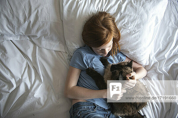 Junges glückliches rothaariges blauäugiges Mädchen auf weißem Bett mit Katze liegend.