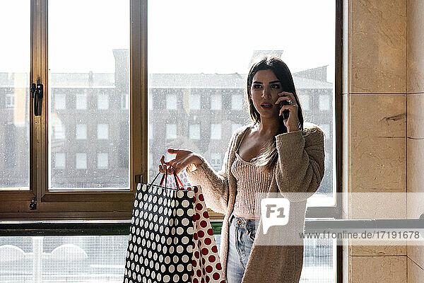 Spanisches brünettes Mädchen  das mit dem Handy telefoniert und Einkaufstüten in einem Wohnhaus hält.