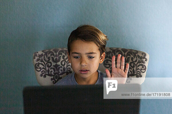 Junge  der an seinem Laptop Schularbeiten erledigt und die Hand zum Sprechen hebt.