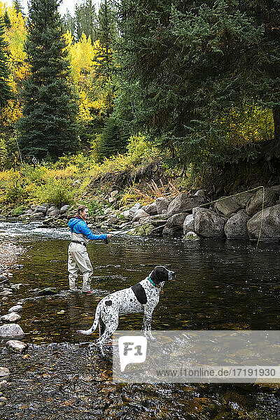 Seitenansicht einer Frau beim Fliegenfischen mit Hund am Flussufer im Wald