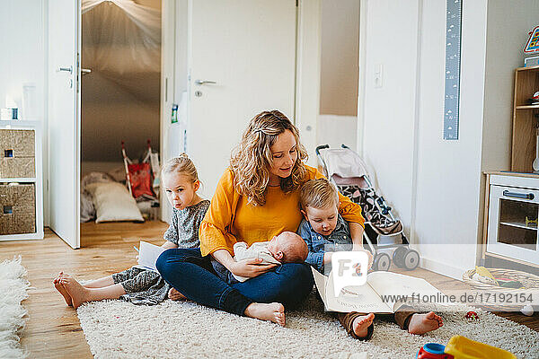 White mother hugging children reading books sitting on rug at home