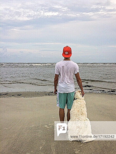 Junger Mann in kurzen Hosen steht mit seinem Hund und schaut auf das Meer hinaus
